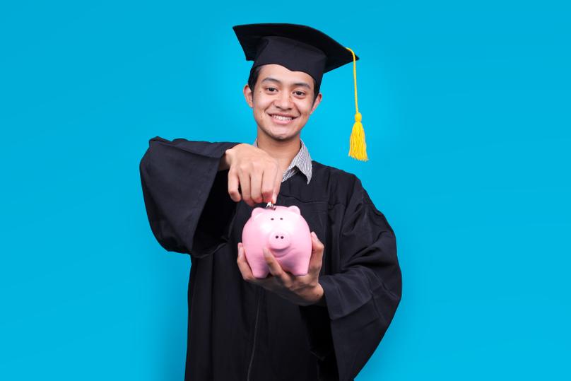 Graduate Man Inserting Coin In Piggy Bank Isolated on Blue Background