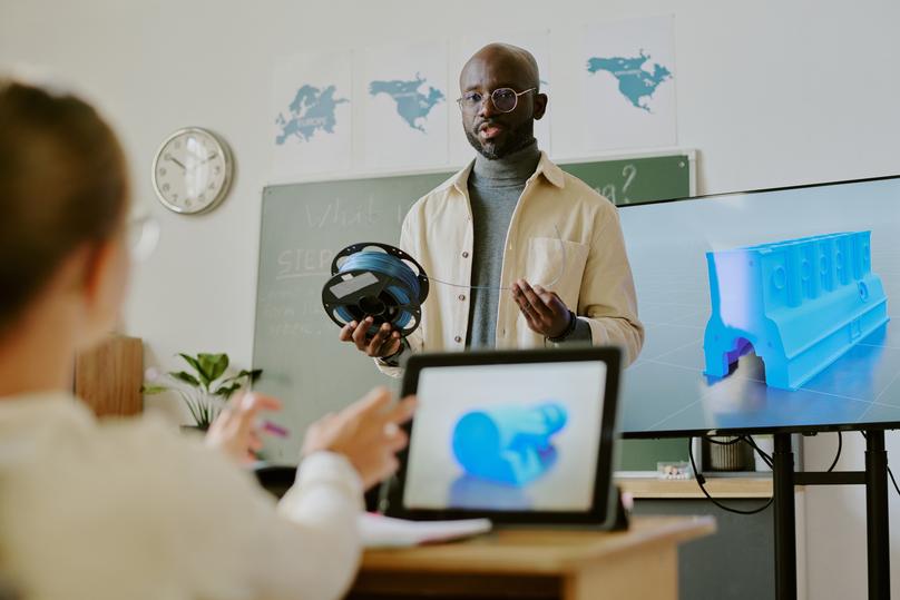 Man presenting virtual reality headset and discussing 3D model displayed on screen in classroom setting. Engaging in interactive learning environment with students observing attentively