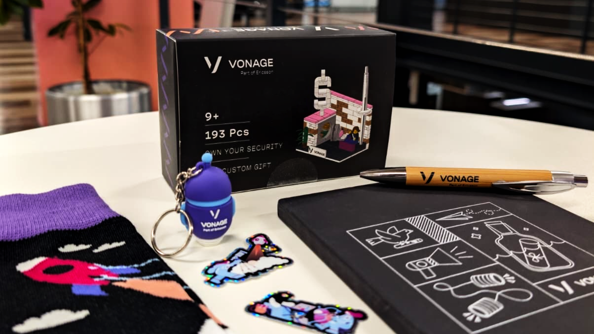 Photo of Vonage-branded merchandise displayed on a round table, featuring a custom 193-piece brick set, illustrated notebook, branded pen, purple socks with a Vonage design, keyring, and colorful character stickers. These items are part of a limited-time giveaway celebrating the launch of the Vonage Developer LinkedIn page.