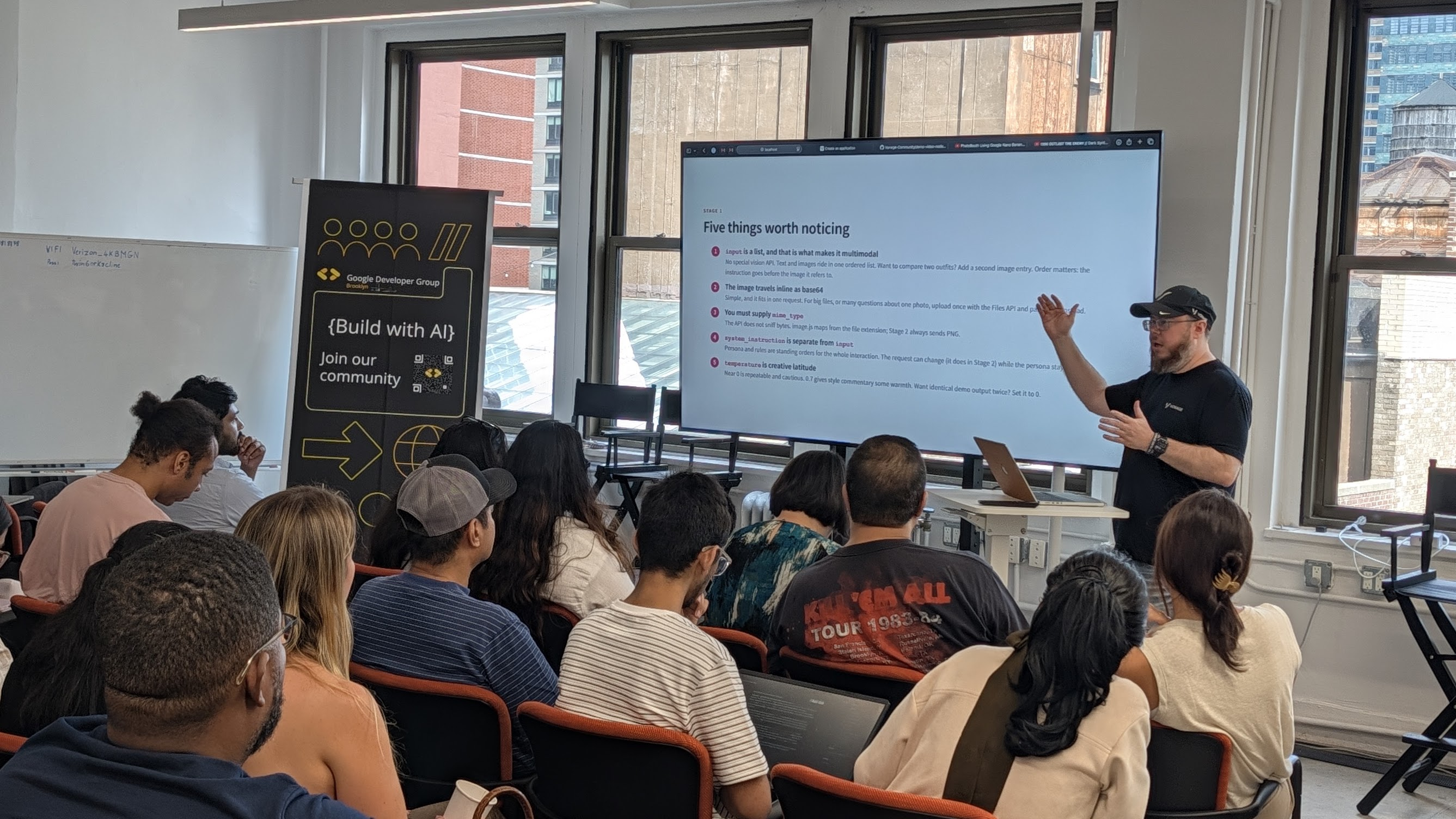 A presenter speaks to an audience in a classroom setting, with "Build with AI" and "Join our community" banners and a screen showing a presentation.