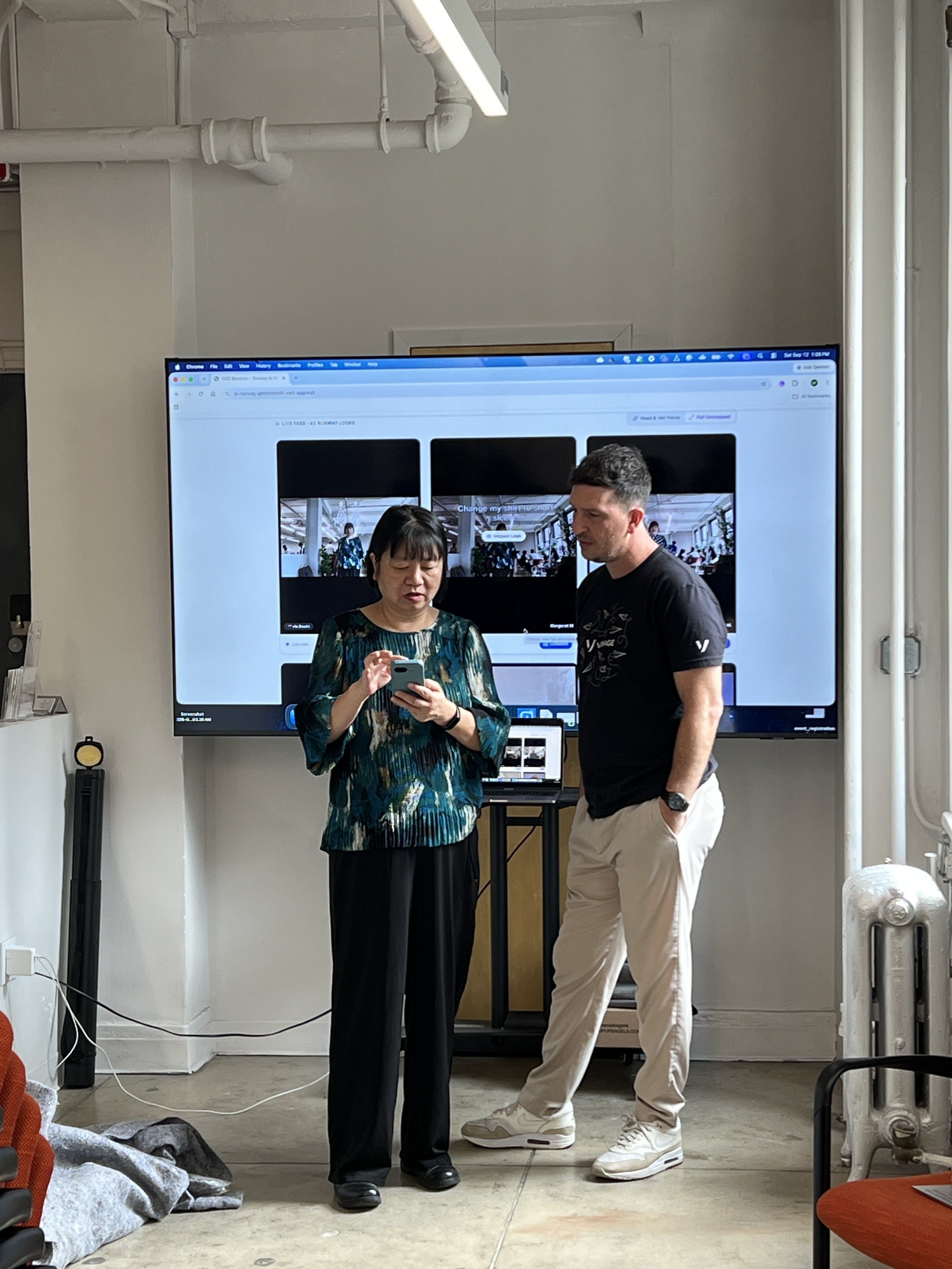Two people stand in front of a large screen displaying images. One is holding a smartphone while the other looks on attentively.