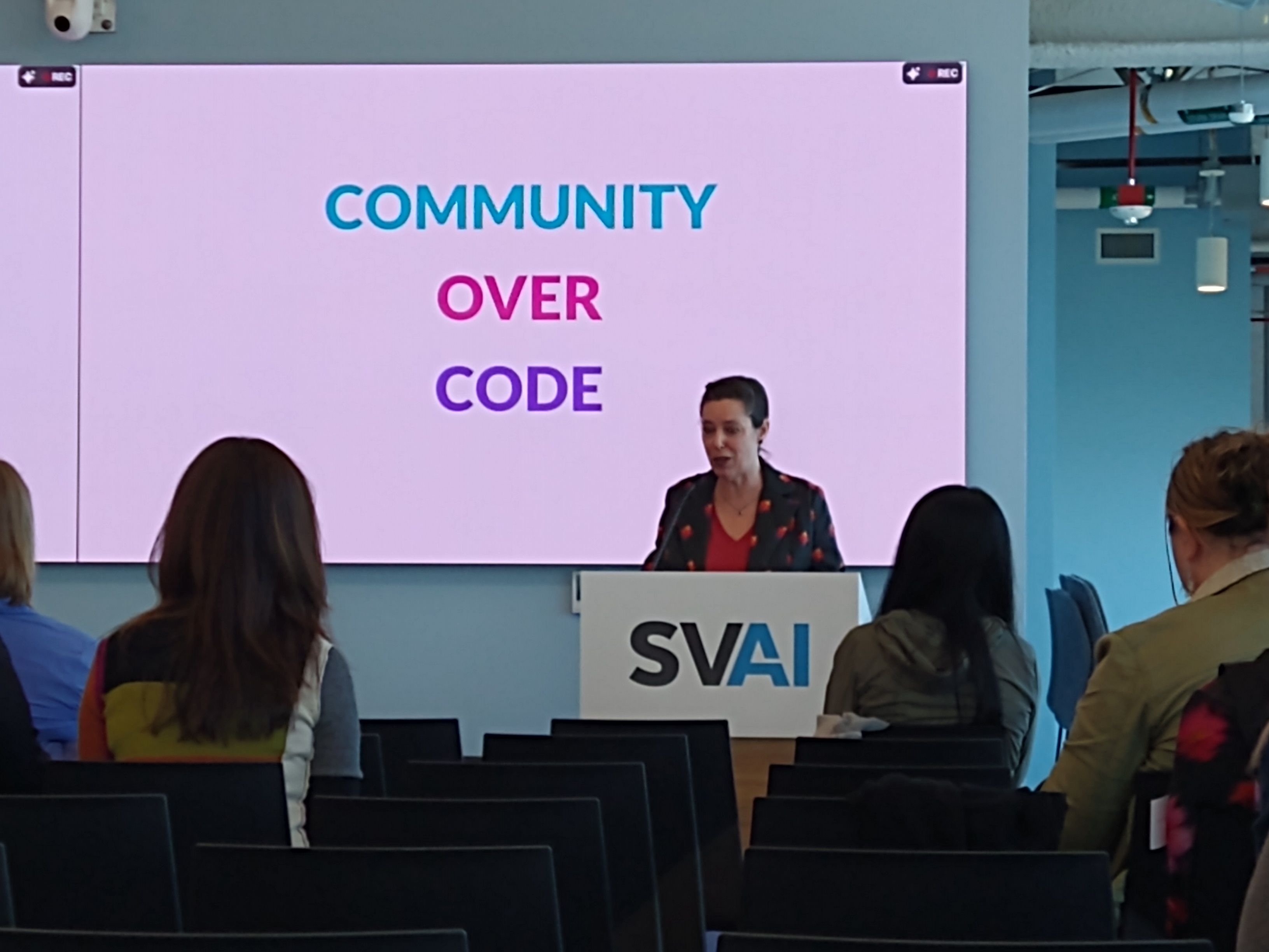 Ruth Suehle emphasizes the role of community in open source at the Women+ in Open Source Day at the SVAI Hub. She is standing at a podium with a slide behind her that says, "Community over code."