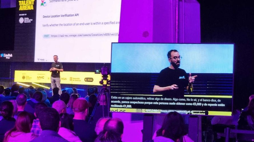 A speaker presents on stage at a tech conference, with a large screen displaying slides and a sign language interpreter visible.