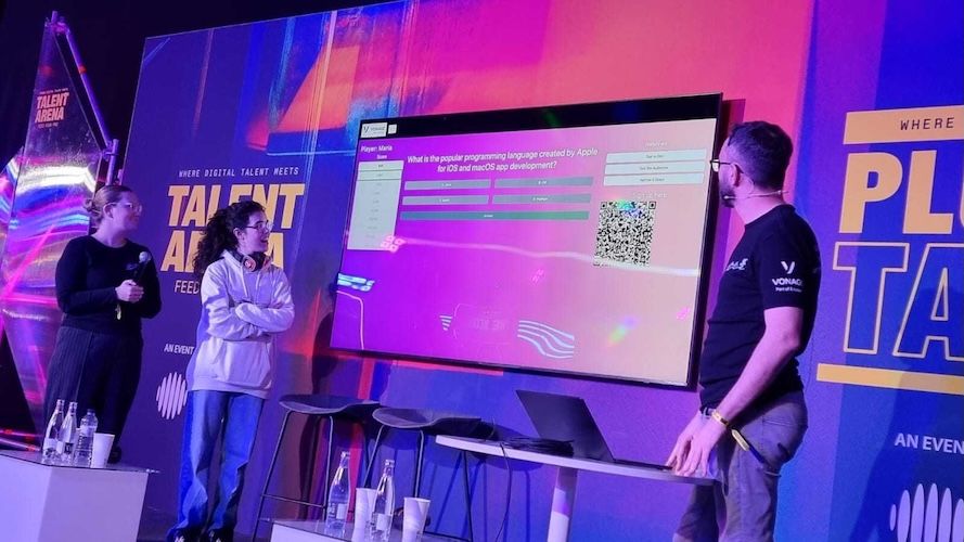 Three people stand on stage at a tech event, discussing a screen displaying a poll about popular programming languages.