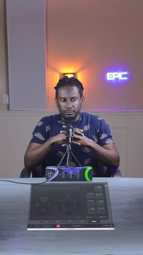 Person sitting at a table with audio equipment, under a neon "EPIC" sign, adjusting a microphone.