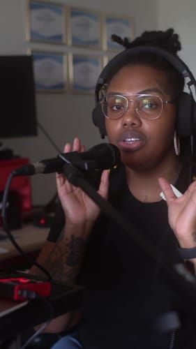 Person with headphones speaking into a microphone, gesturing with hands, in a room with framed certificates on the wall.