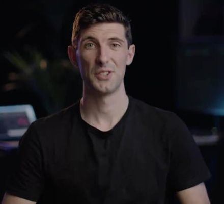 A person in a dark shirt sits in a dimly lit room with audio equipment and a laptop in the background.