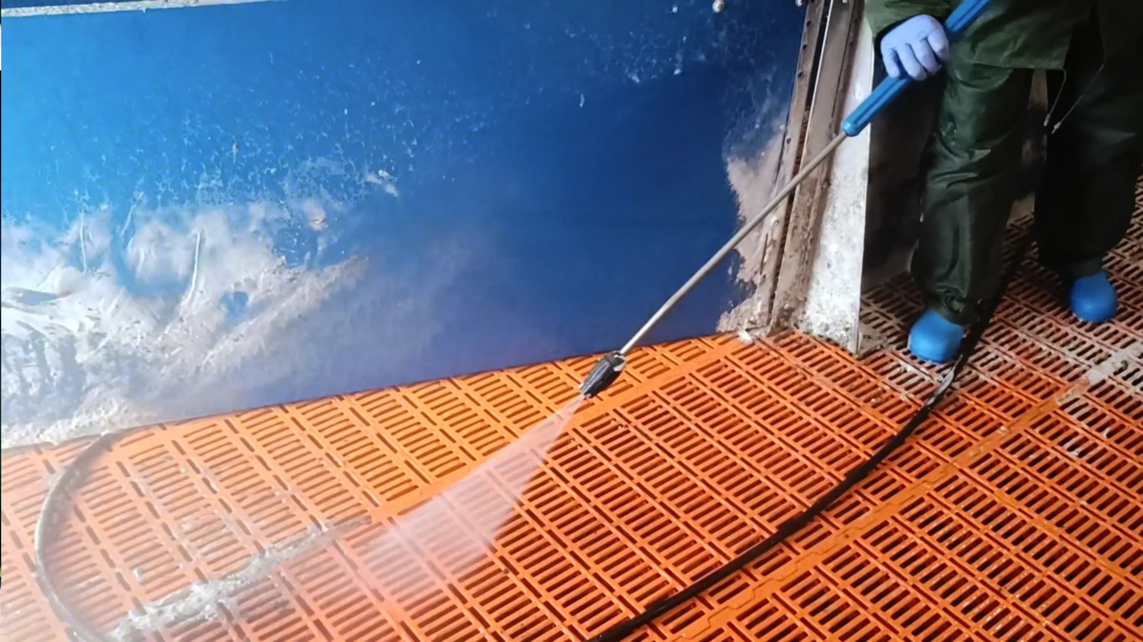 Person cleaning an orange grid floor with a high-pressure water hose, wearing blue gloves and boots.