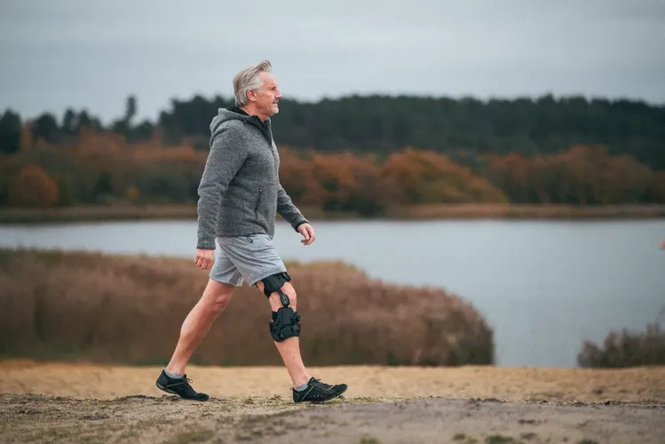 Älterer Mann mit Knieorthese läuft in Natur, im Hintergrund See und Bäume