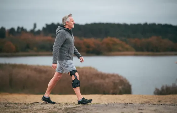 Älterer Mann mit Knieorthese läuft in Natur, im Hintergrund See und Bäume