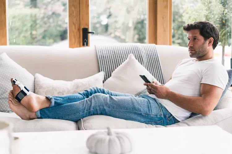 Mann mit Hallux Valgus liegt mit hochgelegten Beinen auf einer Couch und tippt aufs Handy, am Fuß eine Zehenorthese