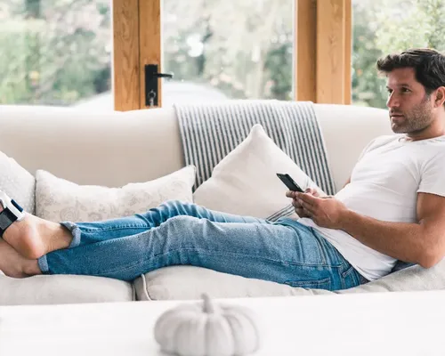 Mann mit Hallux Valgus liegt mit hochgelegten Beinen auf einer Couch und tippt aufs Handy, am Fuß eine Zehenorthese