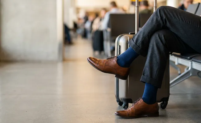 Beine eines Mannes im Anzug am Flughafen; blaue Kompressionsstrümpfe sind unter seiner Hose sichtbar; neben ihm ein kleiner Koffer