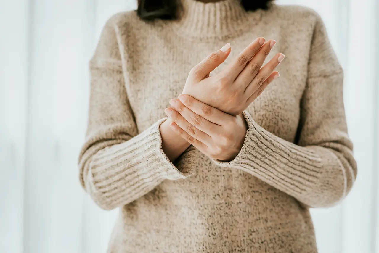 Frau hält ihre Hand wegen Arthrose im Handgelenk, braucht Schiene oder Bandage, trägt beigen Wollpullover