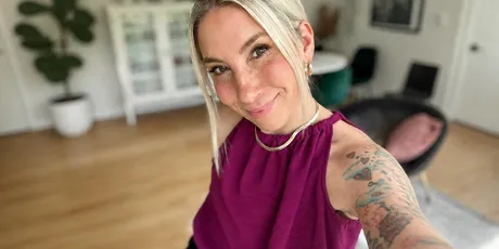 A person with a tattooed arm and blonde hair smiles while taking a selfie in a modern, well-lit room with plants and stylish decor.