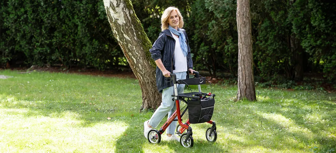 Frau mit Parkinson mit Rollator auf grüner Wiese, im Hintergrund Bäume, Charcot-Marie-Tooth-Syndrom