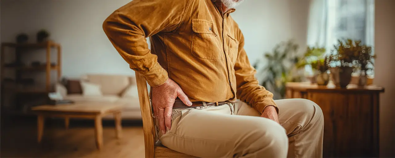 Älterer Mann sitzt auf einem Stuhl im Wohnzimmer und fasst sich schmerzverzerrt an die Hüfte