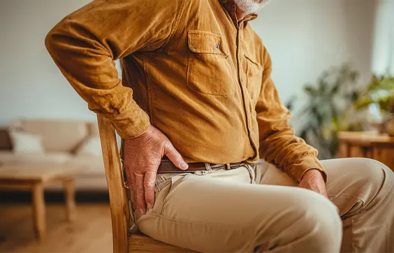 Älterer Mann sitzt auf einem Stuhl im Wohnzimmer und fasst sich schmerzverzerrt an die Hüfte