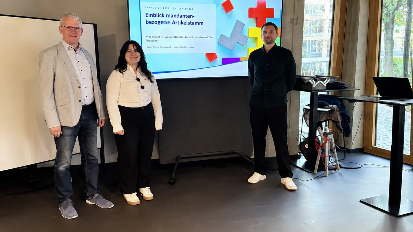 Three people standing in a conference room, with a slide presentation on a digital screen and a whiteboard beside them.