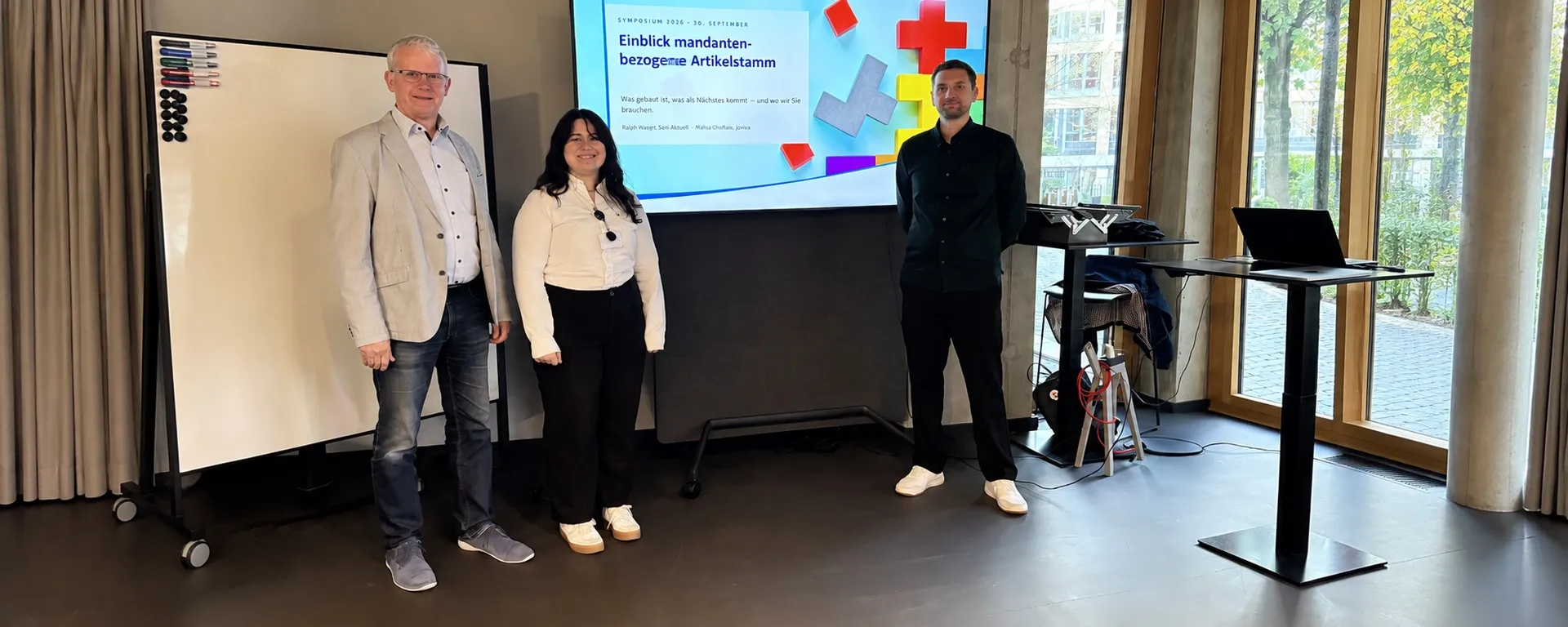 Three people standing in a conference room, with a slide presentation on a digital screen and a whiteboard beside them.
