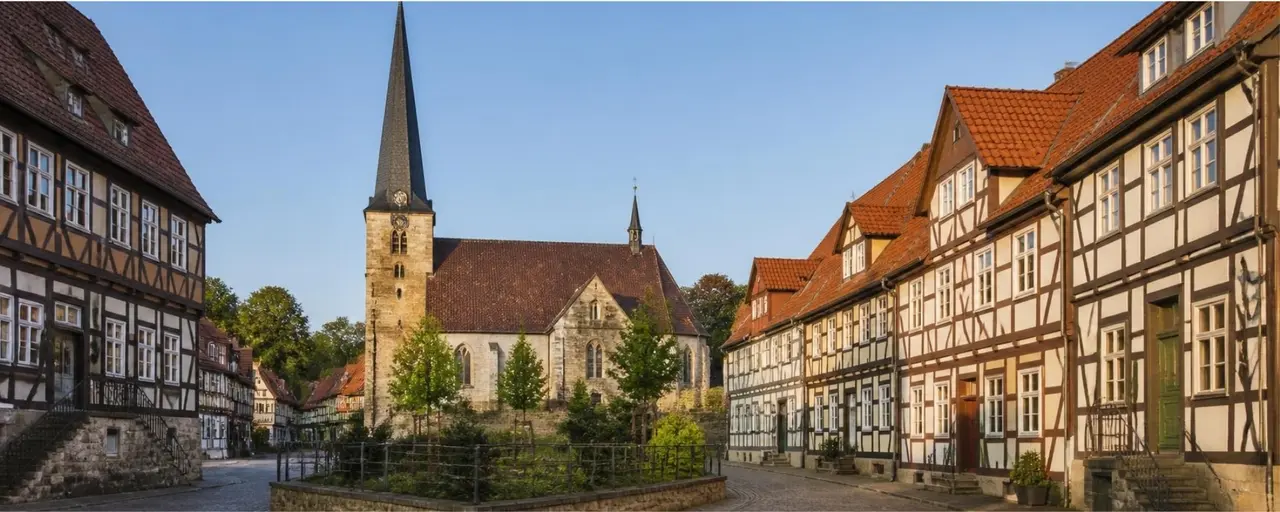 A picturesque European village square with half-timbered houses and a stone church with a tall spire under a clear blue sky.