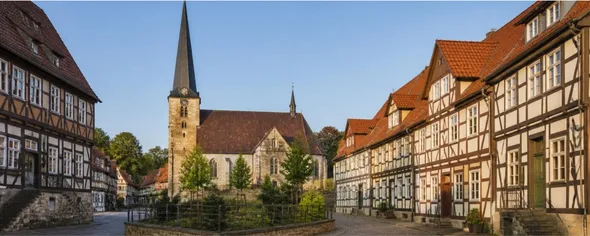 A picturesque European village square with half-timbered houses and a stone church with a tall spire under a clear blue sky.