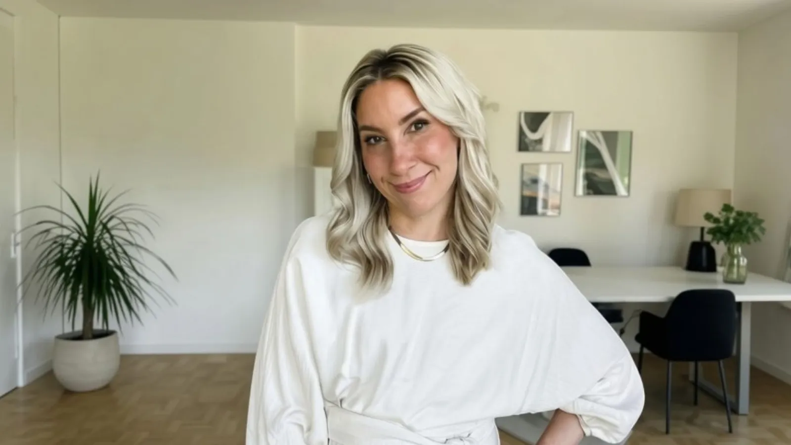 A woman with blonde hair smiles in a modern, bright living room with plants, a white table, and minimalistic decor.