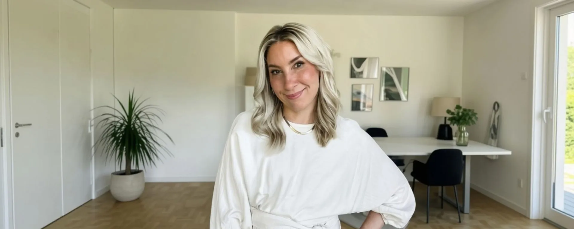 A woman with blonde hair smiles in a modern, bright living room with plants, a white table, and minimalistic decor.