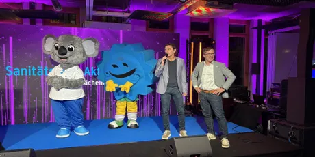 Two men onstage with a koala and star-shaped mascot at an event, featuring a purple-lit background and text behind them.