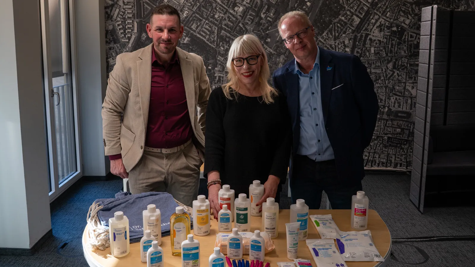 joviva-Mitarbeiterin Karin Pütz mit zwei Mitarbeitern der Dr. Schumacher GmbH in einem modernen Bürogebäude vor einem Tisch mit Hygieneprodukten