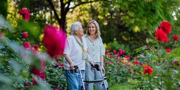 ältere Frau läuft mit Rollator, junge Frau daneben, am Arm eingehakt, gehen spazieren durch Blumengarten, lächeln