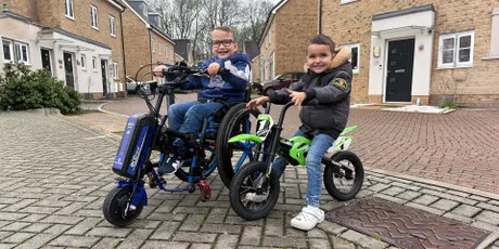 Zwei lachende Jungen auf einer Wohnstraße: Der ältere sitzt in einem elektrischen Kinderrollstuhl mit Zugaufsatz, der jüngere sitzt auf einem grünen Laufrad daneben.