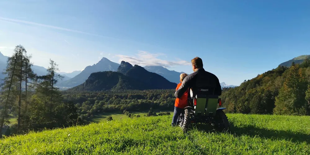 Mann im Rollstuhl umarmt kleinen blonden Jungen vor einer grünen Berglandschaft