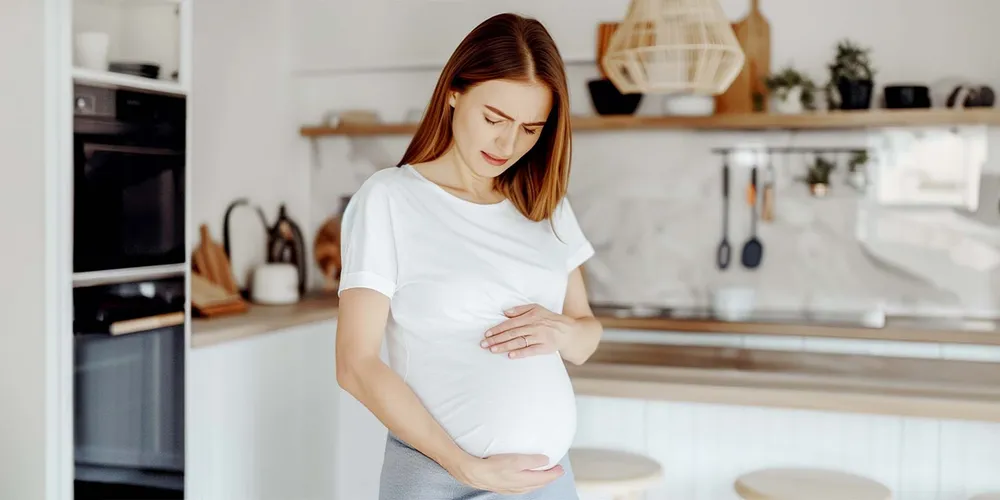 Schwangere Frau mit Inkontinenz hält sich den Bauch, gequälter Gesichtsausdruck, steht in Küche