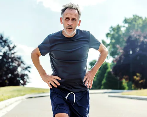 Mann mit Schleimbeutelentzündung hält sich die Hüfte beim Joggen, Weg, Wiese und Bäume im Hintergrund