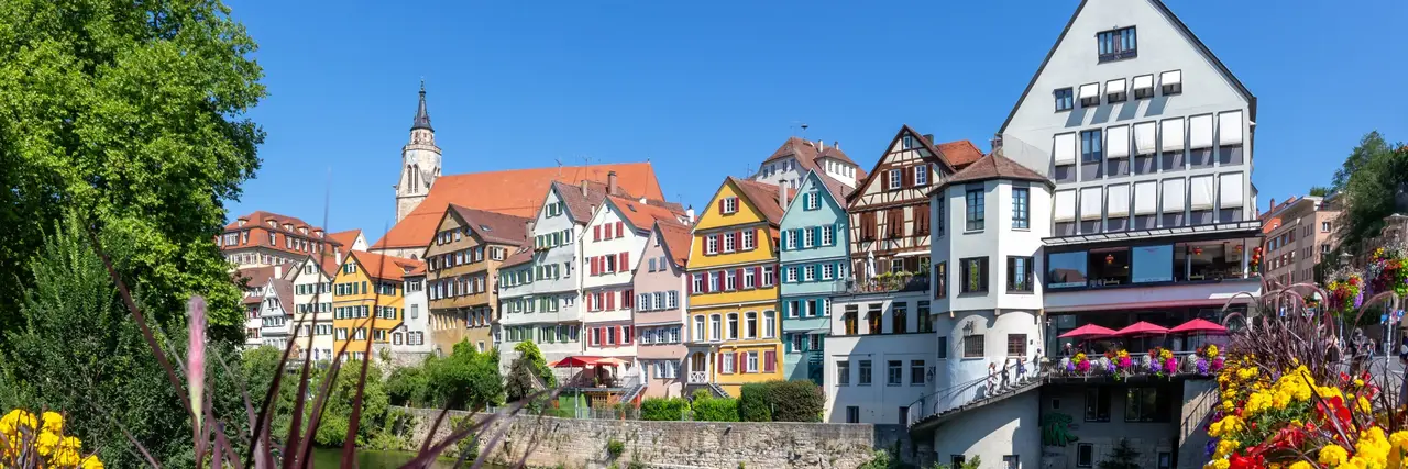 Unter strahlend blauem Himmel säumen farbenfrohe historische Gebäude das Flussufer, im Vordergrund leuchten bunte Blumen, und in der Ferne ist ein Kirchturm zu sehen.