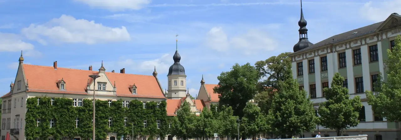 Historische Gebäude mit roten Ziegeldächern und einem Turm, umgeben von Grün, unter einem blauen Himmel mit Wolken.