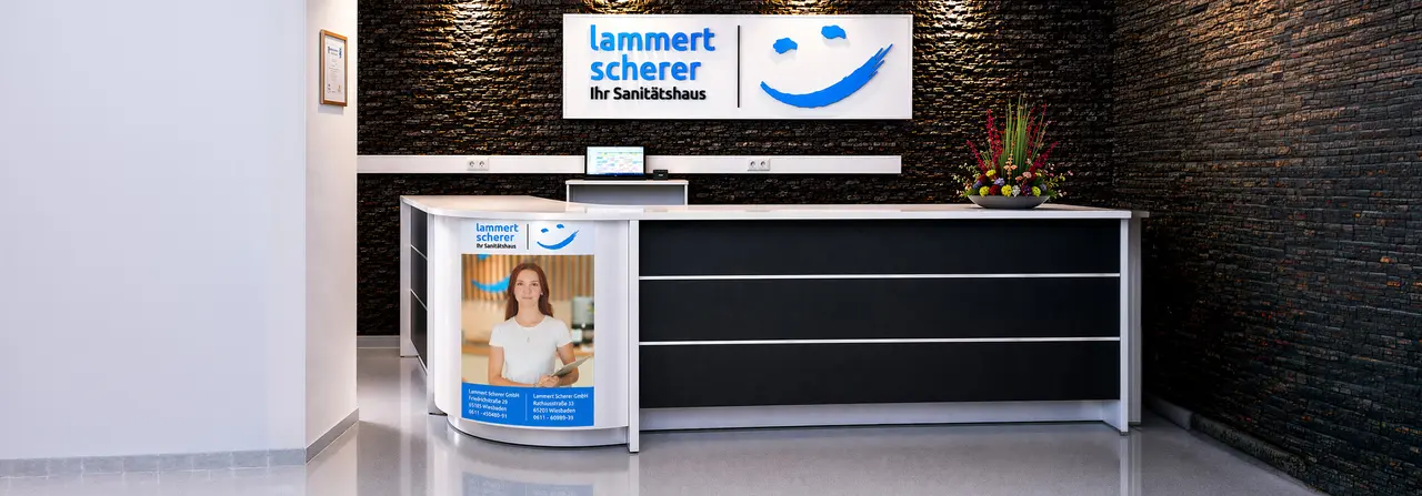 Innenansicht des Empfangs des Sanitätshauses Lammert Scherer GmbH in Wiesbaden; Theke mit schwarzer Front vor einer dunklen Steinwand; im Hintergrund das Smiley-Logo von Sanitätshaus Aktuell und "Lammert Scherer Ihr Sanitätshaus"