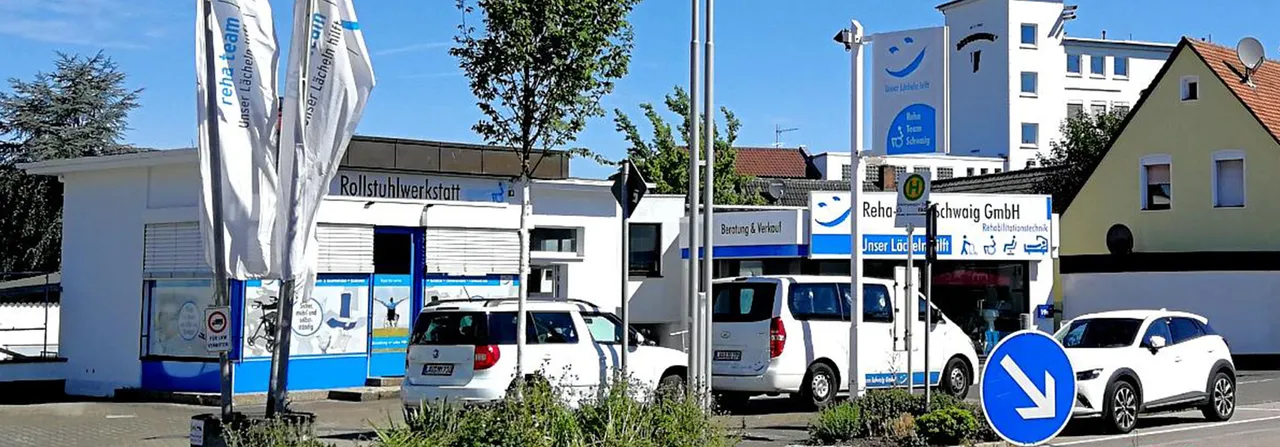 Außenansicht der Reha-Team Schwaig GmbH in Schwaig bei Nürnberg mit weißer Fassade, Rollstuhlwerkstatt, Beratung & Verkauf sowie Firmenfahrzeugen auf dem Parkplatz.