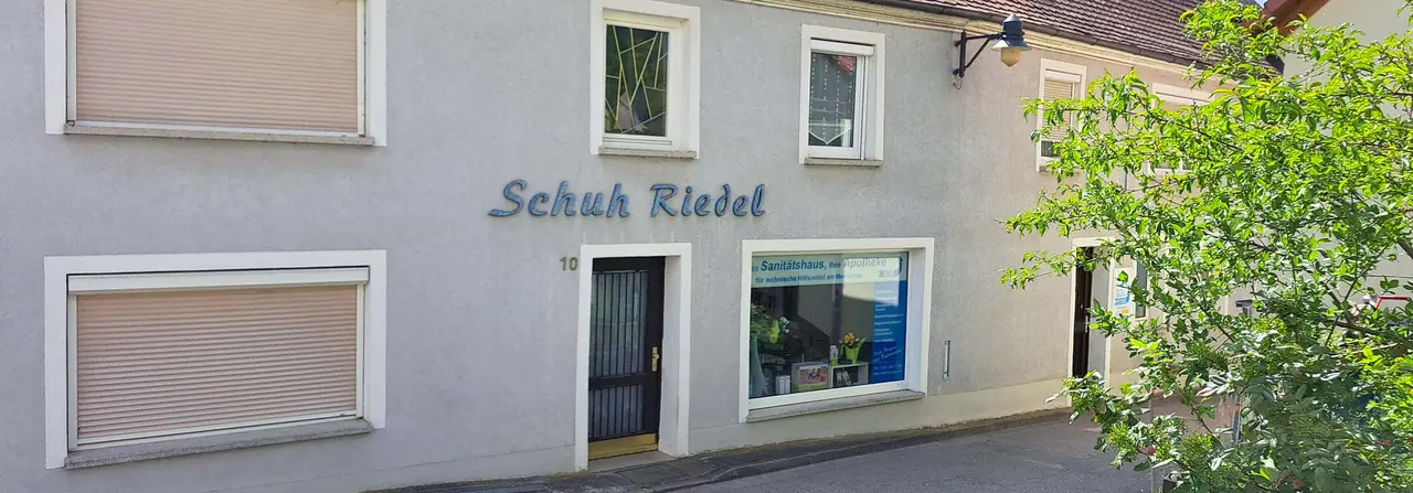 Außenansicht des Sanitätshauses Riedel Pfeuffer in Pleinfeld, untergebracht in einem hellen Wohnhaus mit Schriftzug „Schuh Riedel" an der Fassade.