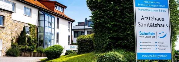 A medical services center with a building featuring a stone and glass façade. A sign reads "Ärztehaus Sanitätshaus" with contact details and services.