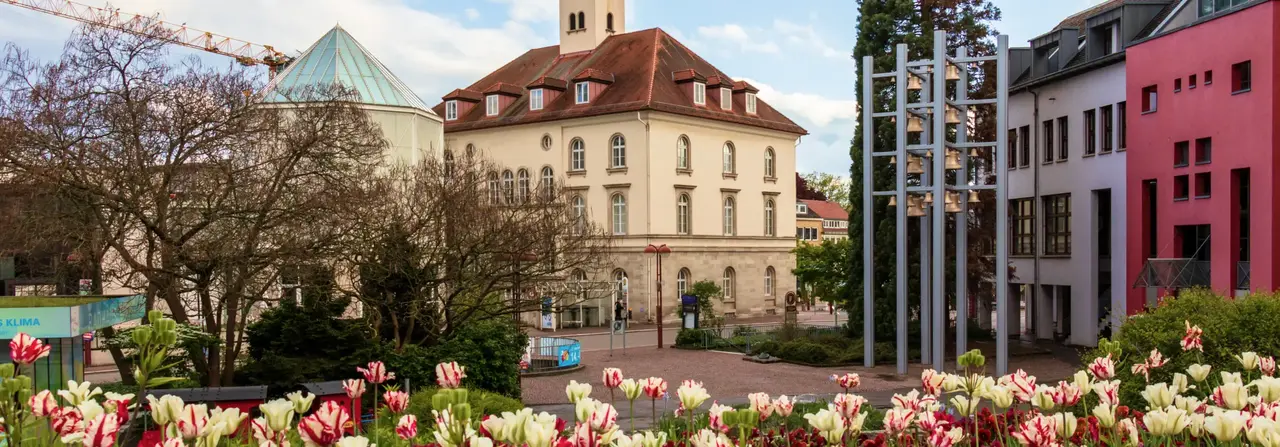 Ein historisches Gebäude mit einem Uhrturm auf einem begrünten Platz, umgeben von Blumen und modernen Gebäuden, unter einem teilweise bewölkten Himmel.