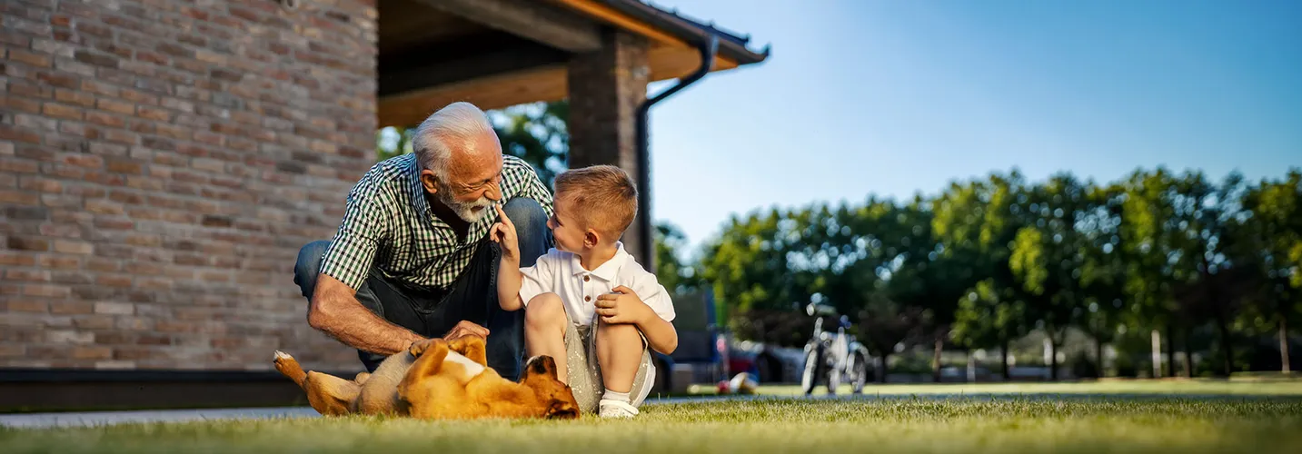 Älterer Mann und kleiner Junge sitzen gemeinsam auf einer Wiese, lachen sich gegenseitig an und streicheln einen Hund.