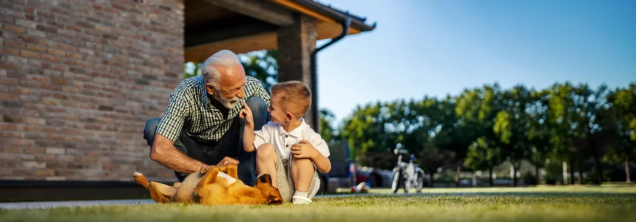 Älterer Mann und kleiner Junge sitzen gemeinsam auf einer Wiese, lachen sich gegenseitig an und streicheln einen Hund.