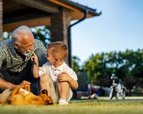 Älterer Mann und kleiner Junge sitzen gemeinsam auf einer Wiese, lachen sich gegenseitig an und streicheln einen Hund.