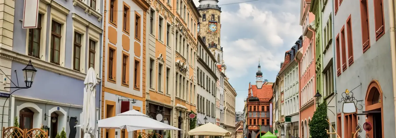 Eine malerische europäische Straße, gesäumt von farbenfrohen historischen Gebäuden, Straßencafés mit Sonnenschirmen und einem Uhrturm im Hintergrund unter bewölktem Himmel.