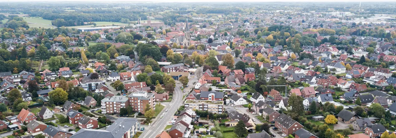 Luftaufnahme einer Vorstadt mit zahlreichen Häusern, von Bäumen gesäumten Straßen und weitläufigen Grünflächen in der Ferne unter einem bewölkten Himmel.