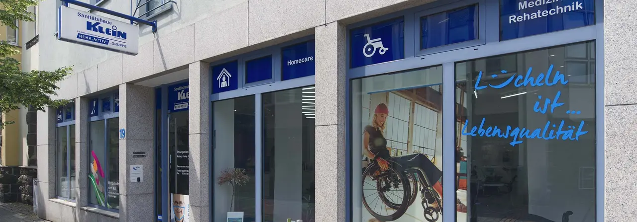 Außenansicht des Sanitätshaus Klein in Gummersbach, einem Ladenlokal mit blau-weißer Fassade. Im Schaufenster ist eine lächelnde Frau im Rollstuhl zu sehen, daneben der Schriftzug „Lächeln ist … Lebensqualität". Über den Eingängen weisen blaue Schilder mit Piktogrammen auf die Bereiche Homecare, Medizintechnik und Rehatechnik hin.