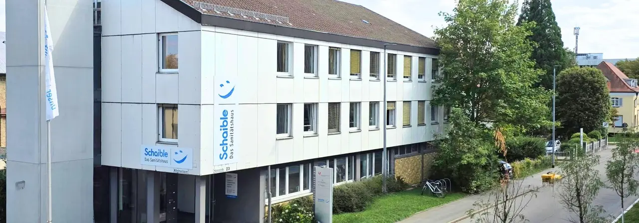 A three-story building with white paneling and several windows, surrounded by greenery. A sign on the building reads "Schaible Das Sanitätshaus."