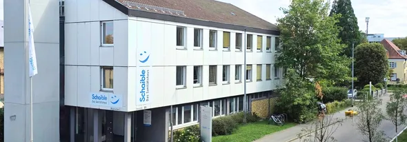 A three-story building with white paneling and several windows, surrounded by greenery. A sign on the building reads "Schaible Das Sanitätshaus."